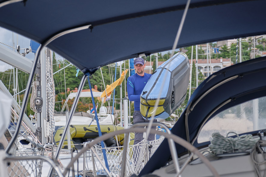 Ein Mann trägt ein blau-grünes Packraft zu einem Segelboot.